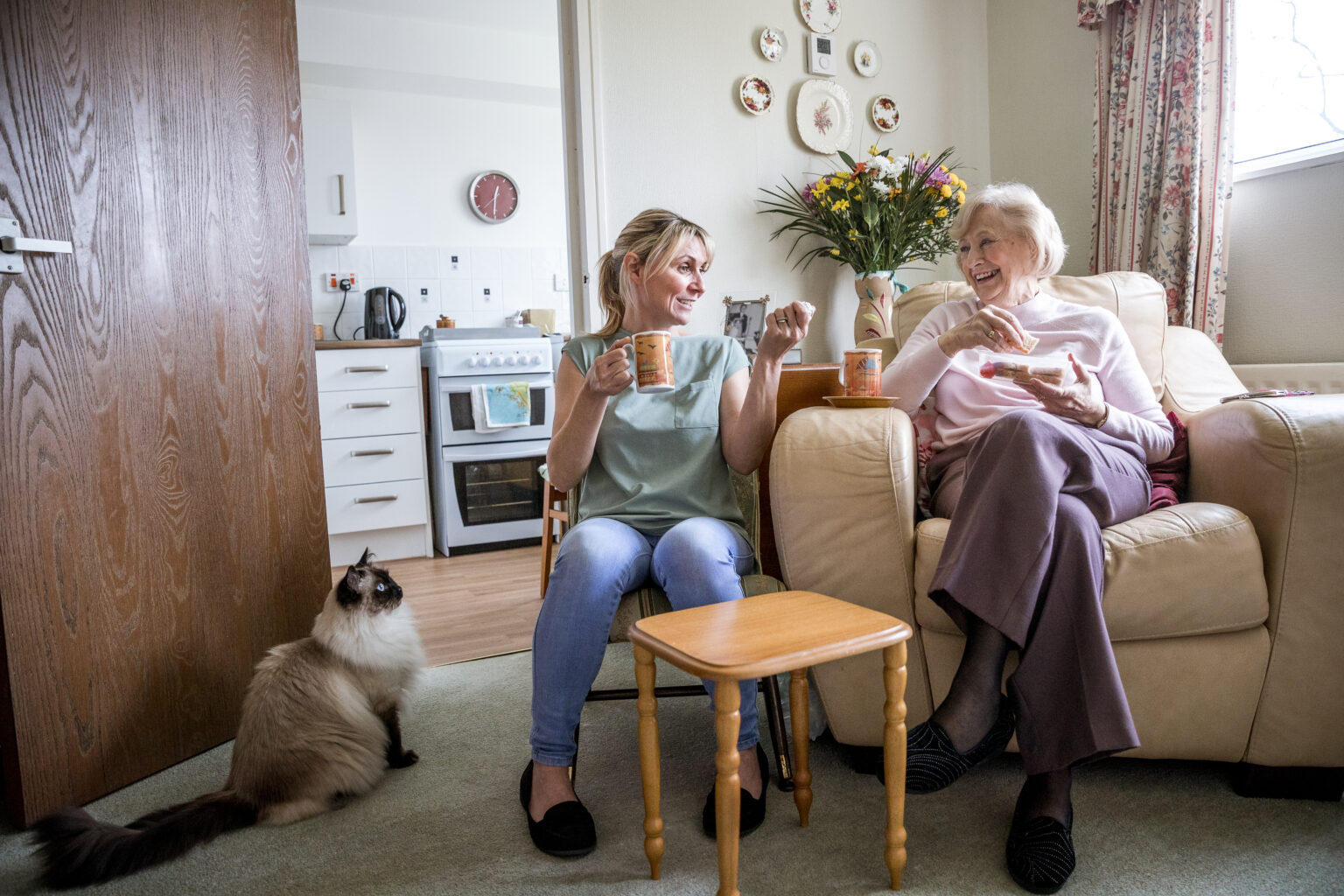 Senior woman sitting in her armchair listening to a younger woman talking. Both look happy as they enjoy a drink and snack together